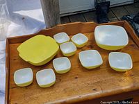 Lot of 10 yellow Pyrex dishes on wooden tray including covered bowl and various sizes of bowls with white interior and yellow exterior.