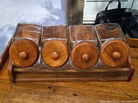 Front view of 4 square glass jars with circular wooden lids in wooden holder