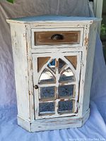 Full view of the whitewashed corner cupboard side table showing front with drawer and glass paneled door.