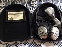 Open hard zip case showing silver Bose QuietComfort 3 headphones with attached cables and a wrapped accessory inside the case pocket. Left side of case has a Bose Courtesy Card in a mesh pocket.