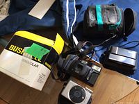 Bushnell binocular box, leather case, Sony Cyber-shot camera and Canon camera on blue fabric, black soft case beside.