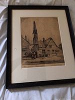 Close-up of a framed sepia drawing of a village scene with church steeple and figures, displaying the signed and numbered print in aged dark wood frame.