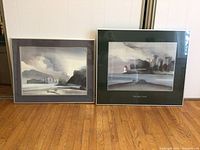 Wide photo showing both framed paintings leaning against the wall on a wood floor, highlighting their size and framing style.