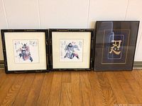 Three framed Asian art prints displayed on wood floor against wall; two with samurai watercolor prints in black bamboo-style frames, one shows Japanese kanji calligraphy in gold on black background in solid black frame.