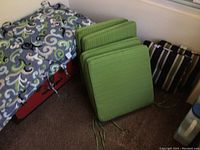 Photo showing four green ribbed square cushions with ties stacked upright beside a large square cushion with a blue, green, white, and black abstract swirl pattern.