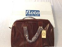 Front view of brown Cenzo leather bag with handles wrapped in plastic, original tag attached, placed in front of Floto box.