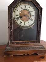 Front view showing the clock face with Roman numerals and central golden dial, framed by carved wooden trim and covered by glass.