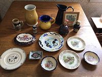 Overview of all ceramic items on a wooden table including pitchers, vases, plates, and smaller dishes showing variety and condition.