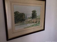 Framed watercolour of a landscape featuring a stone bridge, trees, and distant buildings including a church steeple, hung on a wall.