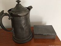 Side view of pewter kettle and rectangular pewter box on wooden surface