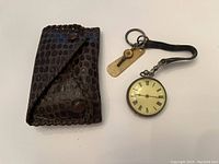 Overall lot including pocket watch with strap and winding key next to leather case, shown on white background.