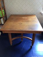 Front view of square wood table showing the tabletop surface with scratches and the leg details with casters.