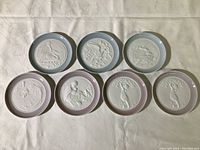 Top-down view of all seven miniature Lladro porcelain plates laid out on a table showing front relief designs and pastel rims, two in light blue and five in lavender.