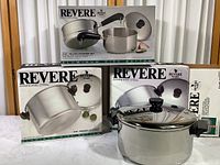 Three boxed new RevereWare stainless steel cookware pieces including stock pot, Dutch oven, and steamer set displayed on table.