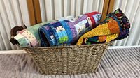 Rectangular wicker basket filled with three rolled handmade patchwork blankets showing colorful fabric patterns and wood handles on basket.