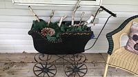 Black antique-style pram with four metal spoked wheels and wooden handles, displayed on a wooden porch floor. Inside the pram are decorative birch logs, green moss, and pine cones, which are not part of the lot.