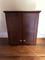 Front view of the closed wooden wall mount medicine cabinet showing two paneled doors with silver knobs.
