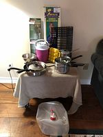 Mixed camping items displayed on a small table, including pans, cooler, grill rack, kettle, egg tray.