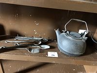 Photo showing cast iron stove top kettle and hanging utensils including ladle, slotted spoon, fork and more arranged on a shelf.