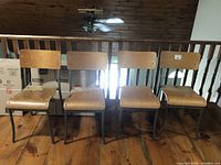 Four stackable chairs with steel frames and wooden seats and backs arranged side by side on a wooden floor in front of a staircase railing.