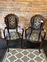 Front view of two matching antique Victorian sitting chairs with carved wood and floral tapestry upholstery, set against brick wall.
