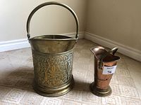 Antique brass coal scuttle with floral embossed design next to antique copper match holder with brass handle and base