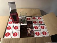 Open box showing multiple unopened boxes with red and white Coca-Cola branding. One glass and one box are positioned on top for display.