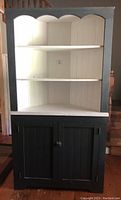Front view of the corner cabinet showing upper white shelves and lower black doors.