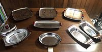 Eight various shaped stainless steel Mid-Century Modern style serving trays arranged on a wooden table, showing overall lot contents and condition.