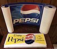 Plastic Pepsi display sign with retro Pepsi-Cola bottle cap and slogan next to two rolls of Pepsi corrugated paper banner showing the modern Pepsi globe logo.