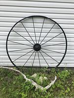 First black antique steel wagon wheel against siding and greenery, showing spokes and central hub detail.