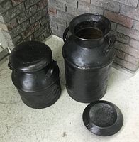 Two large black painted milk cans; one with lid off and placed on ground, one without lid.