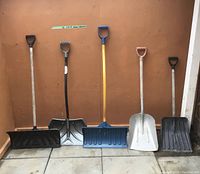 Seven snow shovels in a row against a brown wall: two black plastic flat shovels with wooden and metal handles, one silver metal scoop shovel with wooden handle, one blue plastic shovel with wooden handle, and one black plastic shovel with wooden handle.