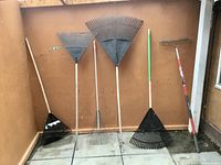 Nine assorted landscaping tools visually arranged against a wooden wall; six rakes with wooden and plastic handles of various types and sizes.