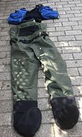Green fly fishing pants laid out showing built-in black neoprene boots and adjustable straps at waist.
