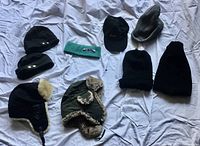 Photo showing 8 men's outdoor headwear items, including toques, balaclava, headband, power beanies, fur-lined winter hats, and a ball cap arranged on a white fabric surface.