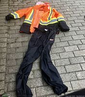 Photo of orange and black men’s rain jacket with reflective stripes paired with black rain pants with zippers at the calf and ankle velcro straps, displayed on brick pavement.