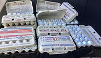 Multiple open cartons filled with white used golf balls, each carton labeled with various brands and size designations, some cartons hold 18 balls, others 12