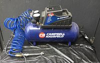 Photo showing blue Campbell Hausfeld air compressor with black motor cover, 110 max PSI label on top, and attached coiled blue air hose with metal air blow gun nozzle.