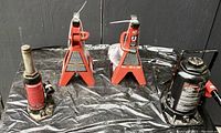 Photo showing two red Motormaster hydraulic bottle jacks and two red Big Red steel car jack stands arranged on a black tarp against a dark background