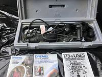 Open gray carrying case with black Dremel rotary tool, attached cord, and black hand grip accessory inside. Three pamphlets laid in front of case including two instruction manuals and one usage guide.