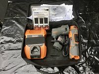 Photo shows all items in the lot inside an open black carrying case on a black surface: cordless orange and gray Rigid drill, orange and gray battery charger, gray Dremel rotary tool with power cord, pack of three Dremel flap sanding belts in original plastic, and instruction manuals.