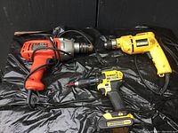 Three drills placed on black surface: one orange Black and Decker corded drill, one yellow DeWalt corded drill, and one yellow/black DeWalt cordless V Max drill with battery pack attached.