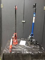 Two heavy-duty utility jacks placed on black table; one red, one blue, both used and with long vertical posts and handles.