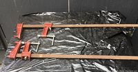 Two large bar clamps laid parallel on black tarp floor, showing orange-red clamping heads and long wooden bars.