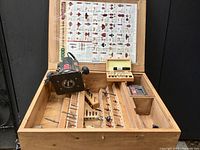 Full view of the Master Grip router placed in a wooden storage box with multiple compartments holding various types of router bits and attachments. A drill bit size chart or router bit identification poster is pinned behind the setup.