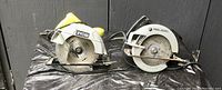 Photo showing two used handheld circular skill saws from Ryobi and Black & Decker placed side by side on a black plastic surface against a black background.