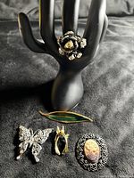 Front view of all five items including butterfly brooch, 3 vintage brooches, and statement ring on a black background and ring display hand.
