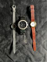 Three wristwatches laid flat on black fabric: center is a black Michael Kors ceramic watch with crystal bezel; right is a Guess watch with brown leather strap and gold-tone case; left is a Guess watch with black leather strap and black case.