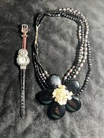 Photo showing the full necklace with multi-strand black onyx and beads along with the large black hibiscus flower pendant featuring white bead cluster at center, shown next to the 1990s ornate silver-tone Brighton style watch with leather strap.